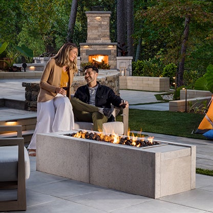 A couple is sitting by an outdoor firepit and smiling with one another